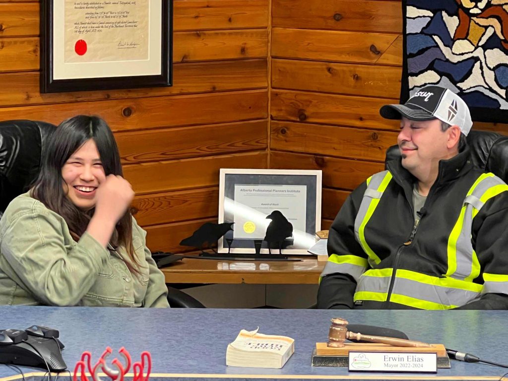 Two people sitting at a table with a book on the table and certificates on the walls behind