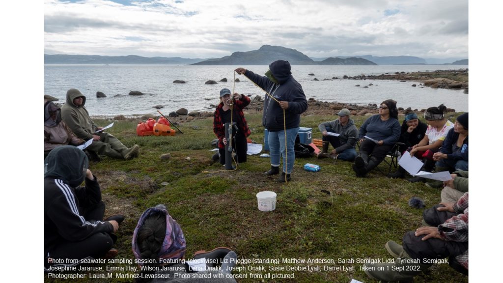 A person demonstrating sampling technique to other participants. Water in the background. Cloudy skies.