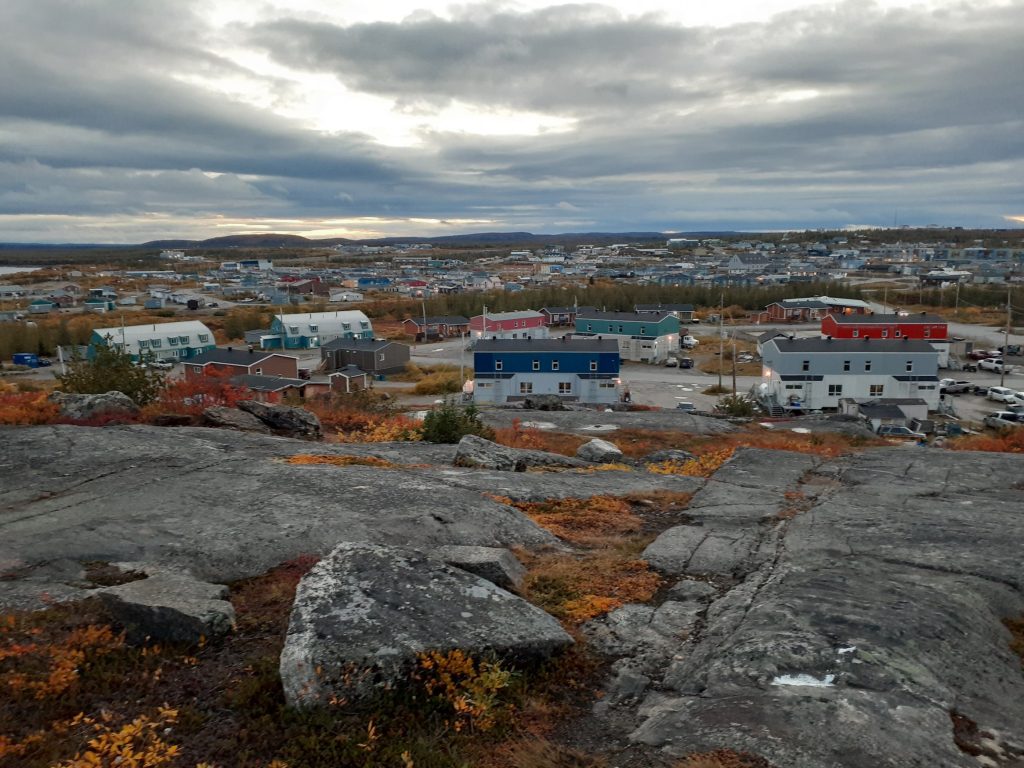 Buildings in the background, rocks with some vegetation in the foreground, under cloudy skies.