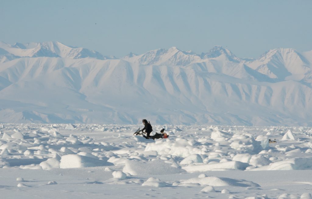 A picture containing a person on a snow scooter with snowy mountains in the background